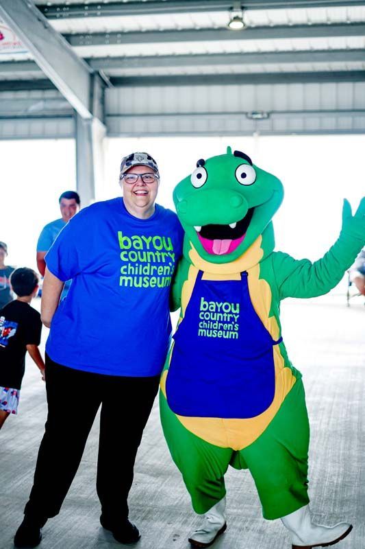 Two people posing indoors, one in a green dinosaur mascot suit, both wearing blue museum shirts.