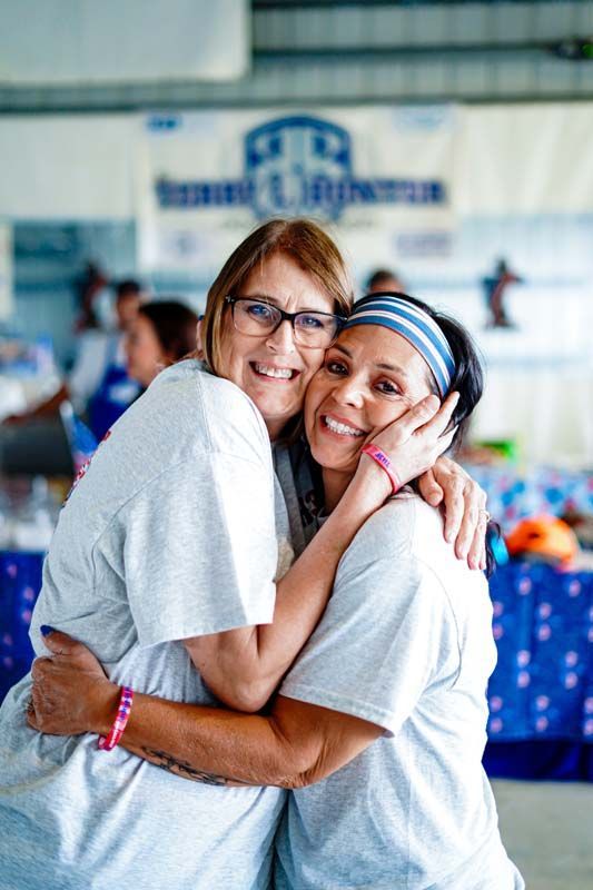 Two people smiling and hugging at an indoor event with a blue tablecloth in the background