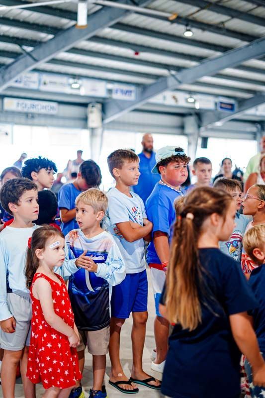 Children gathered in a covered sports pavilion, some in blue shirts and one girl in a red polka-dot dress