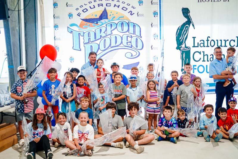 Group of children and adults posing in front of a “Trophy Dodd” event banner, with balloons and sponsor signs
