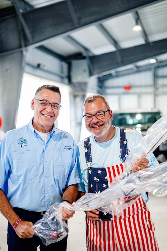 Two smiling men in a warehouse unwrap a large clear-wrapped item together.