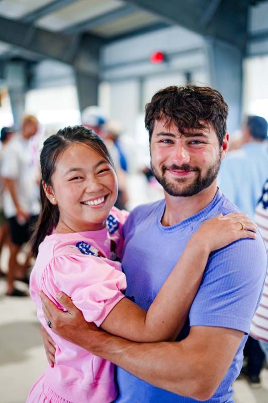 Two people smiling and hugging in a bright indoor space, with others blurred in the background.