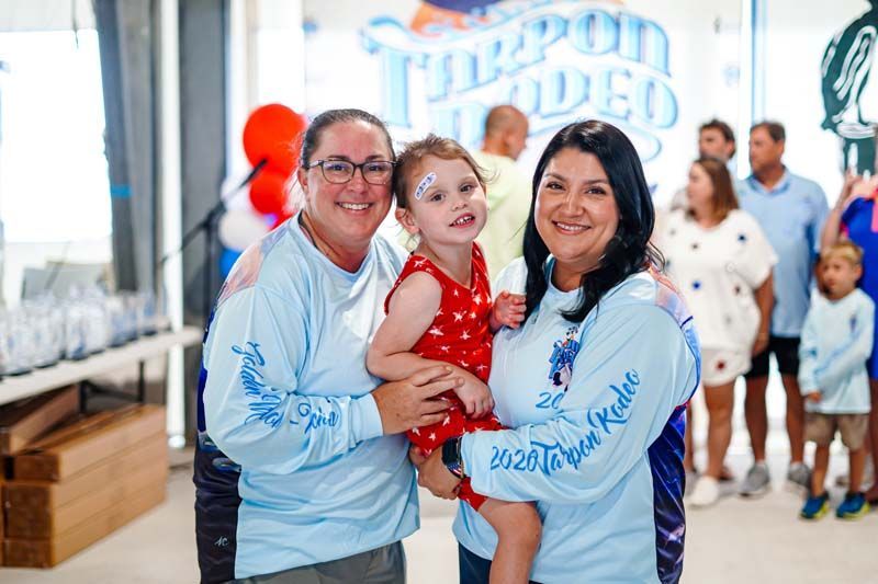 Three people in matching white shirts pose indoors, smiling, with a child in a red dress in the center.