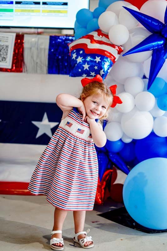 Child in red, white, and blue dress posing beside patriotic balloon decorations and a large star balloon
