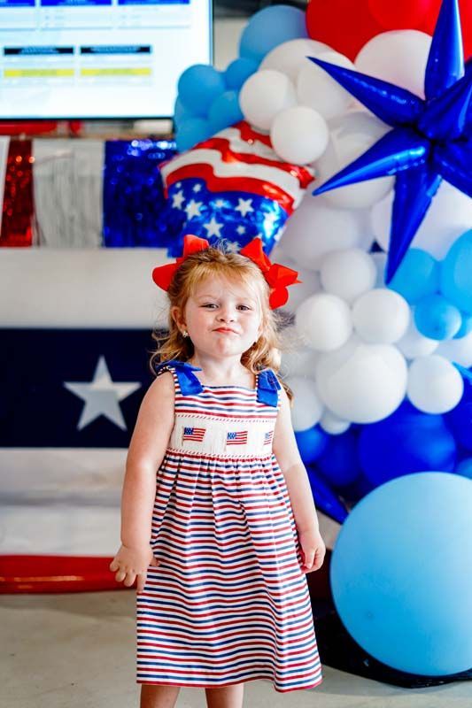 Child in red-and-white dress standing by red, white, and blue balloons at a festive patriotic event