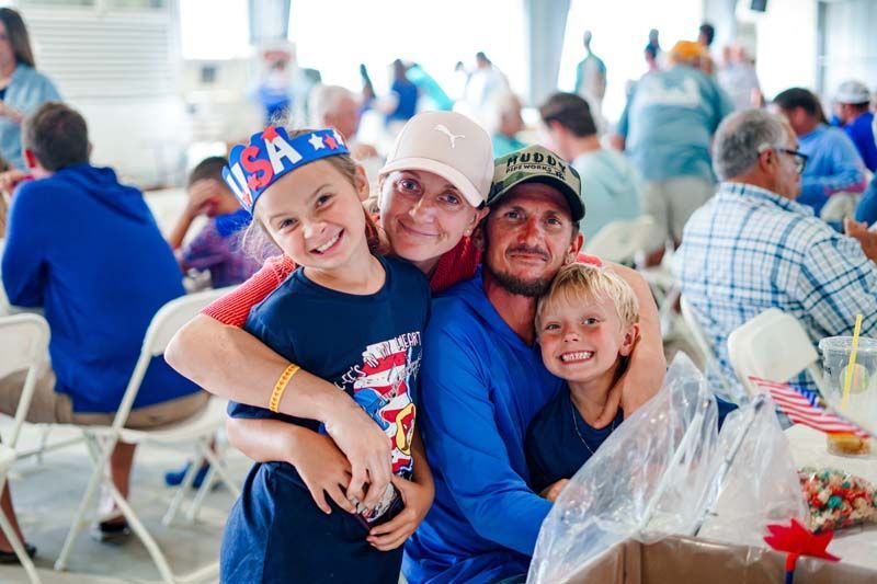 Smiling family in blue shirts and hats posing at a crowded indoor event with plastic bagged gifts on the table