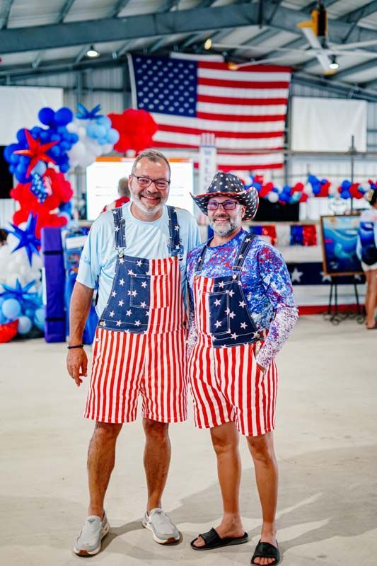 Two smiling people in patriotic aprons pose in front of a large American flag and balloon decorations.