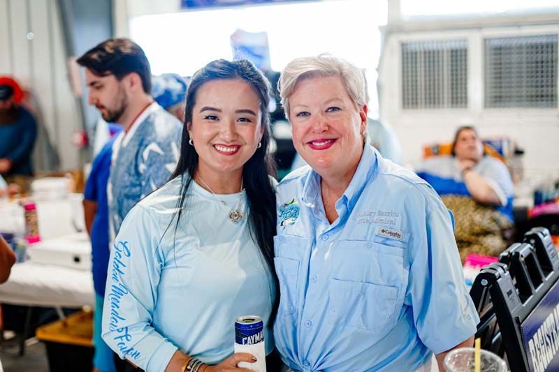 Two smiling volunteers in light blue shirts at a busy indoor event booth