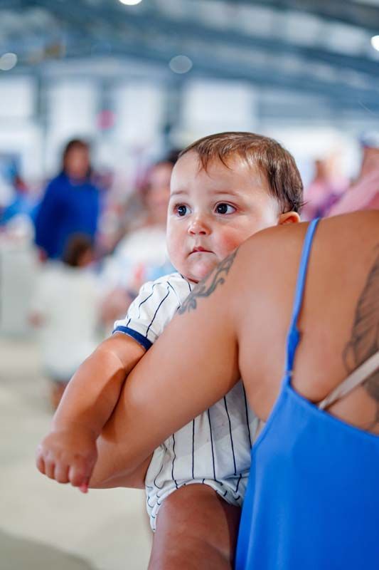 Baby in striped onesie being carried by a woman in a blue top at a busy indoor venue