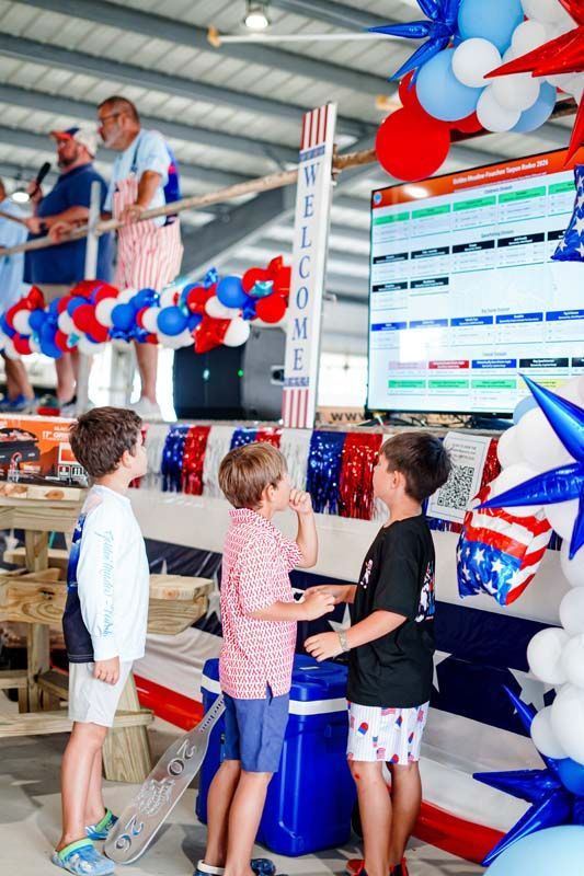 People at a red, white, and blue concession stand with a menu board and balloons.
