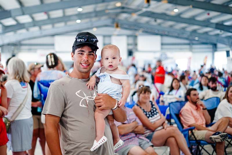 Man holding a baby in a crowded indoor event space, with seated people in the background