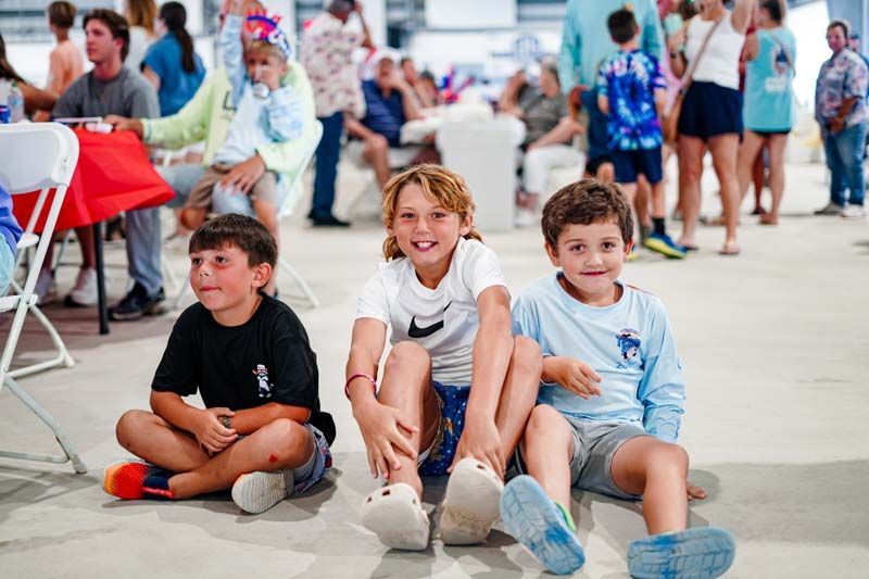 Children sitting on the floor at a crowded outdoor event, smiling and relaxing in casual clothes