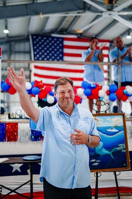 Man waving at a patriotic event with U.S. flag, red-white-blue decorations, and a fish painting behind him