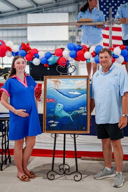 Two people posing beside a framed shark painting at a patriotic outdoor event