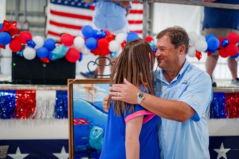 Man in light blue shirt hugs a person at a patriotic celebration with red, white, and blue balloons.