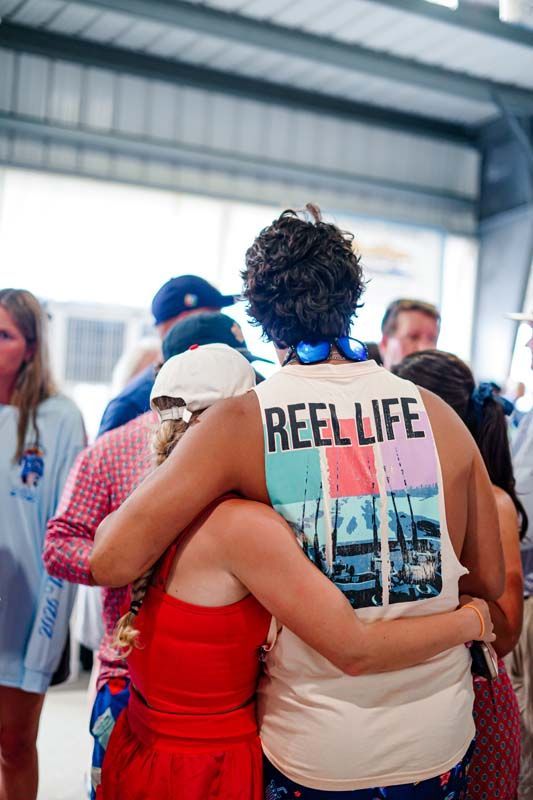 Two people embracing in a crowded indoor event, one wearing a red dress and white cap, the other a beige shirt reading “REEL LIFE.”