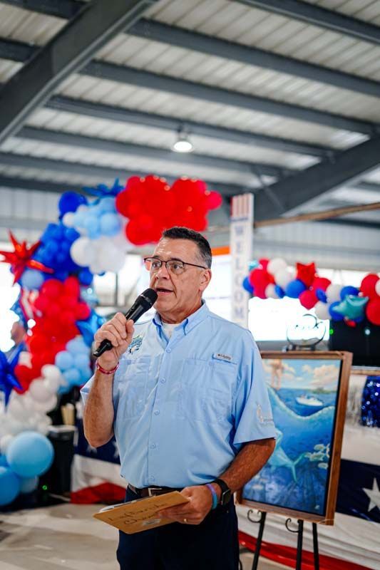 Man speaking into a microphone at a decorated indoor event with red, white, and blue balloons and artwork on display