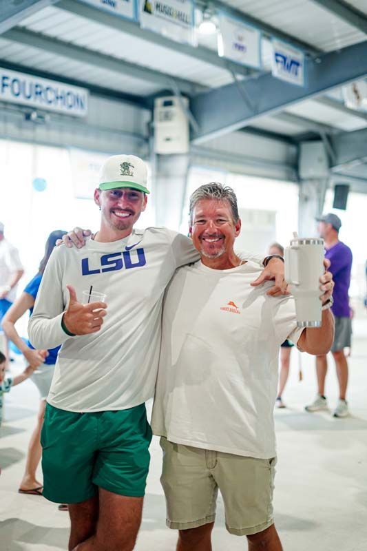 Two smiling men with arms around each other at an indoor event, one in an LSU shirt and cap.