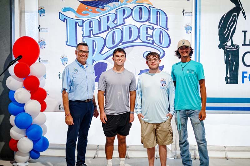 Four men posing on stage at a Tarpon Rodeo event, with red, white, and blue balloons on the left.