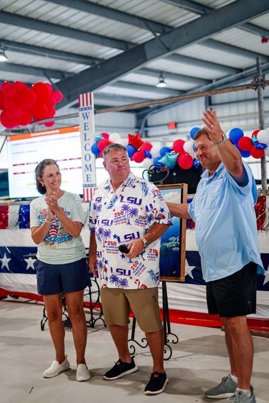 Three people at a patriotic event, one speaking at a podium with red, white, and blue decorations.