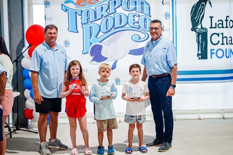 Four people stand with balloons and a trophy backdrop at a Tarpon Rodeo event.