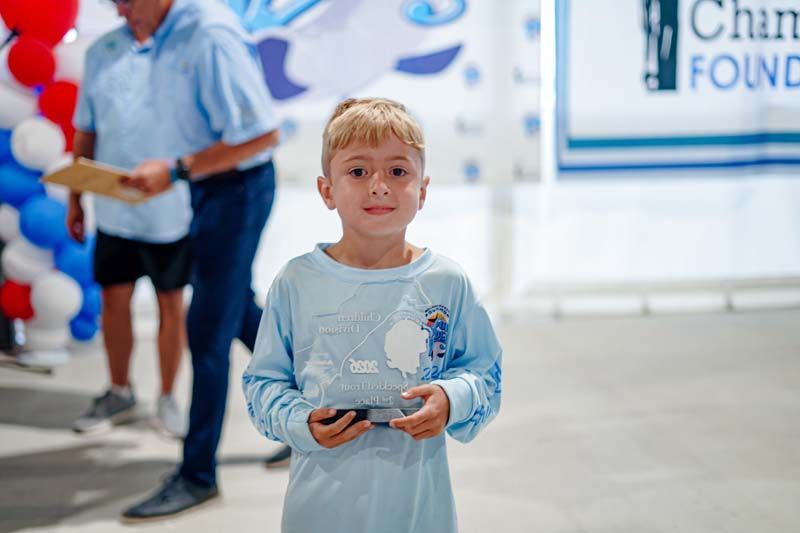 Young child in a light blue shirt holding a phone at a charity event, with people and a banner in the background