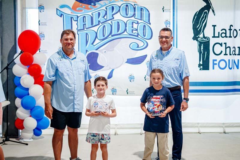 Two men and two children posing in front of a Tarpon Rodeo sign and balloon arch