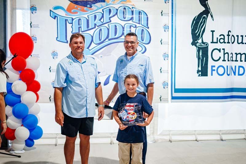 Three people posing at a Tarpon Lodge event with red, white, and blue balloons and sponsor signage
