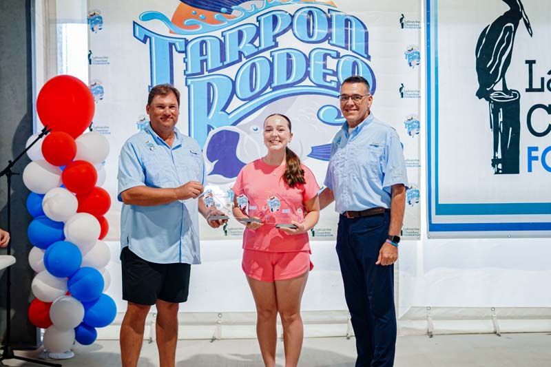 Three people pose at a ribbon-cutting in front of a Tarpon Lodge sign and red, white, and blue balloons.