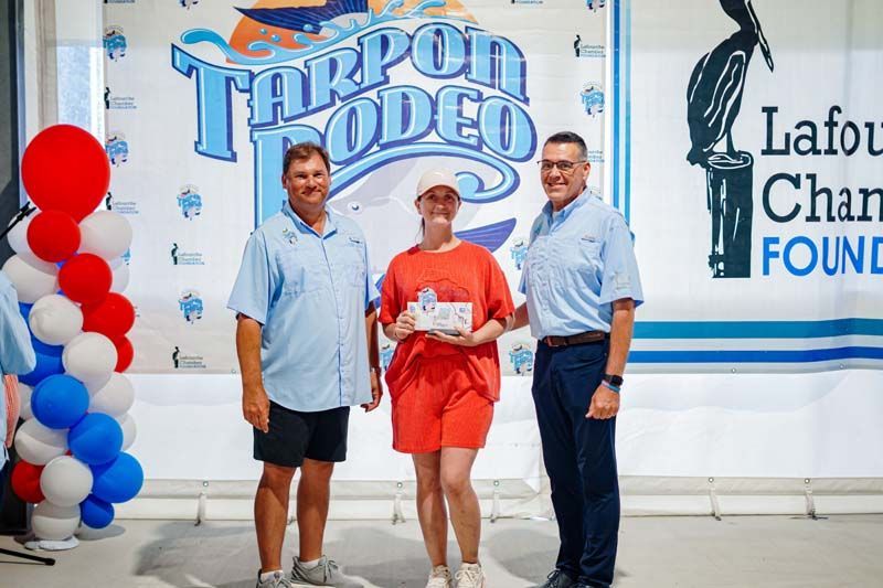 Three people posing at a Tarpon Rodeo event backdrop, with red, white, and blue balloons.