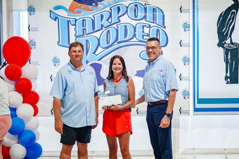 Three people stand in front of a Tarpon Rodeo sign, holding a plaque, with red and blue balloons nearby.