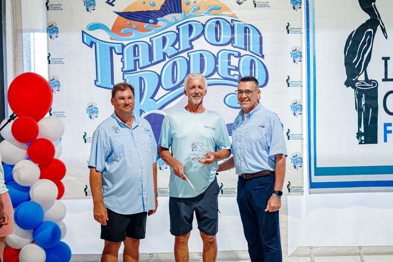 Three men stand in front of a “Therpodrode” banner, holding a plaque at a formal event.