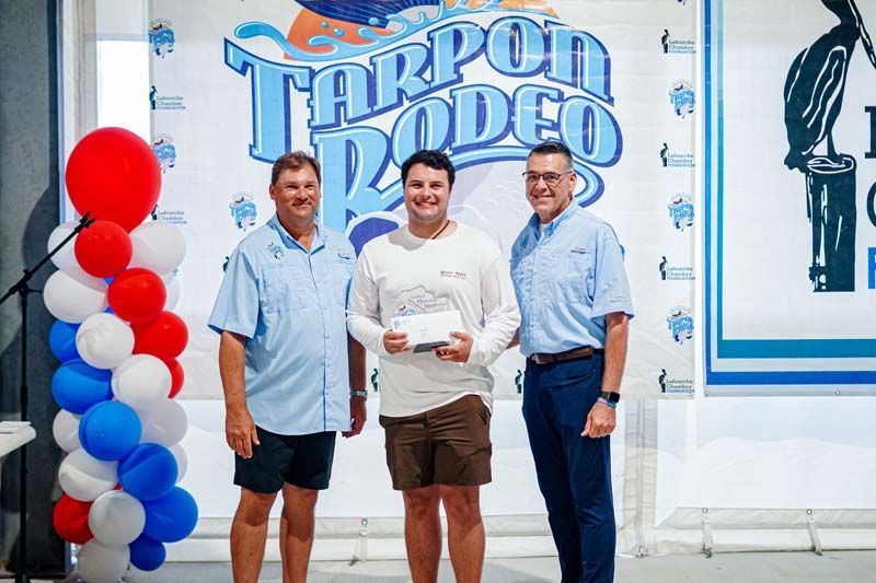 Three men pose with a plaque before a Tarpon Rodeo sign and red, white, and blue balloons.