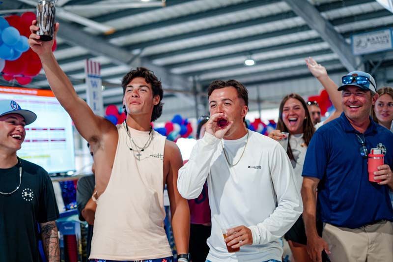 Fans cheering at an indoor event, one holding a trophy and another speaking into a microphone