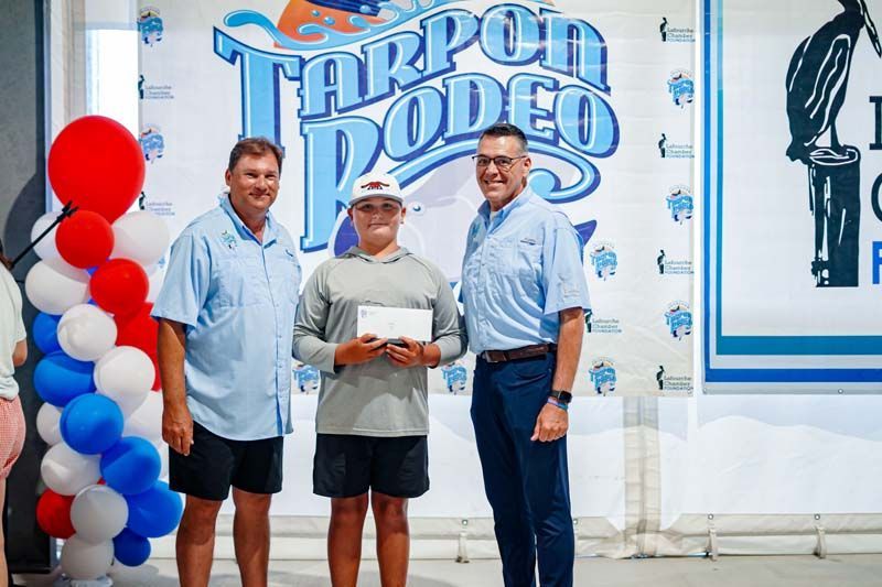 Three men posing in front of a Tarpon Rodeo banner, one holding a plaque, with red, white, and blue balloons.