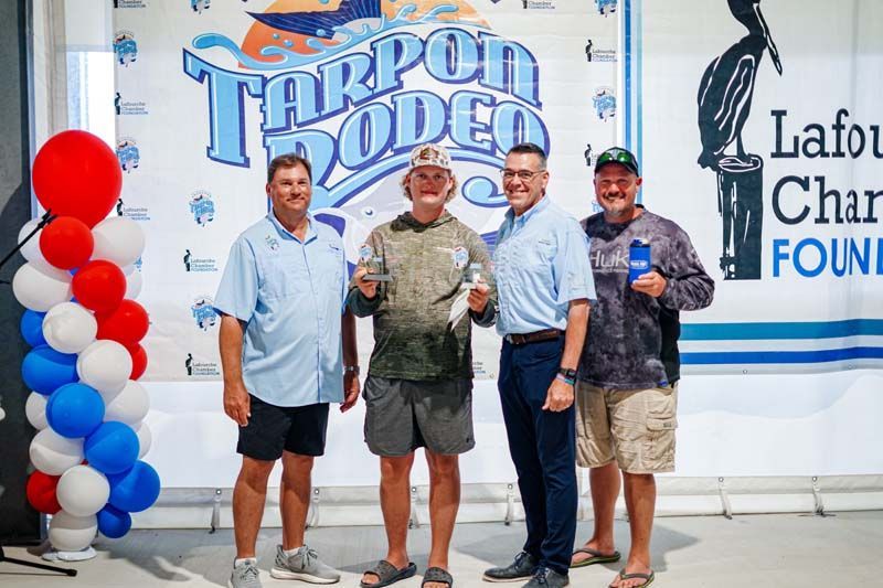 Four men pose on stage before a Tarpon Rodeo and Lafourche Charters Foundation banner, with red-white-blue balloons.