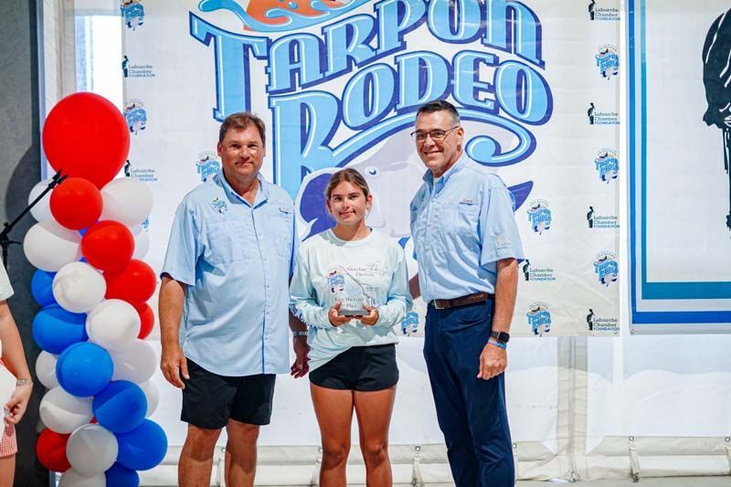 Three people posing with a trophy at the Tarpon Rodeo event, with red and white balloons nearby.