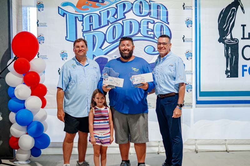 Three people posing with a child at a Tarpon Rodeo event banner, holding a certificate