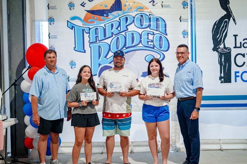 Four people hold trophies at the Tarpon Rodeo wall, with a host standing beside them.