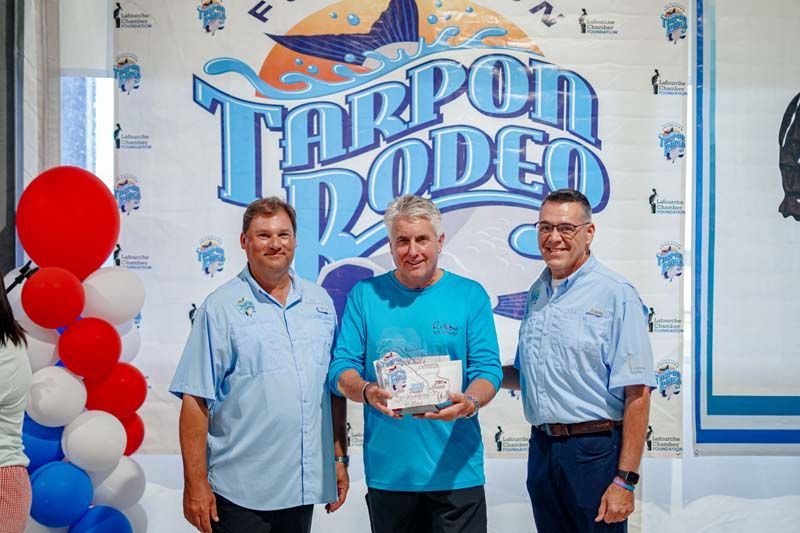 Three men stand in front of a Tarpon Rodeo banner, one holding a plaque, with red and white balloons nearby.