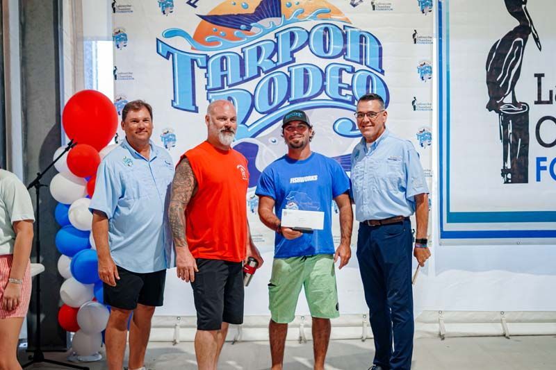 Four men pose at a podium event; one holds a certificate, with red balloons and a “Tarpon Rodeo” backdrop.
