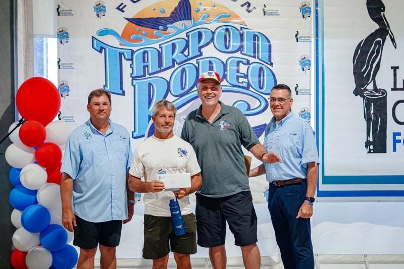Four men pose holding an award in front of a “Tarpon Rodeo” banner and red-white balloons.