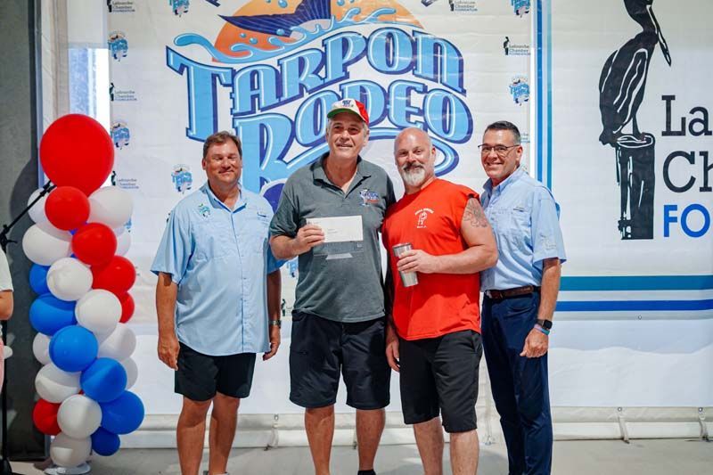 Four men posing in front of a Tarpon Rodeo backdrop, with red, white, and blue balloon columns nearby.