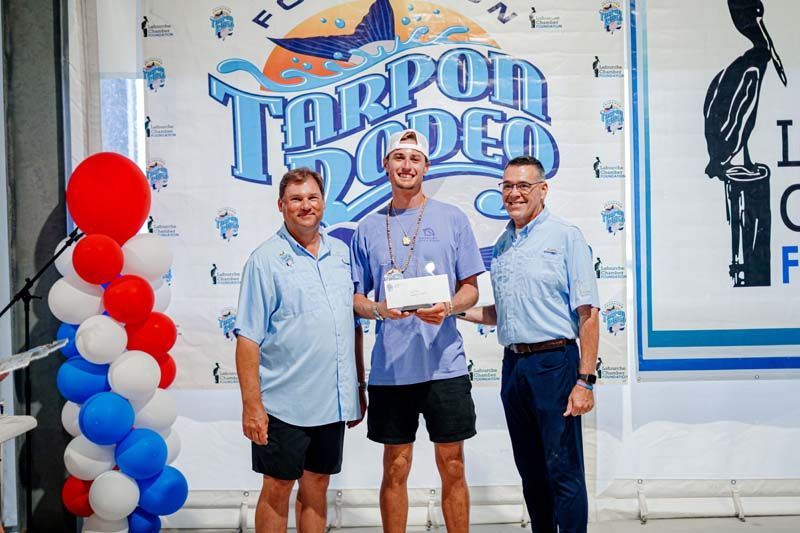 Three men in blue shirts pose with an award plaque at a Tarpon Rodeo event backdrop.