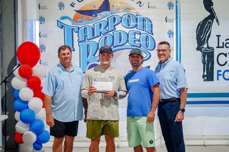 Four men posing at a fishing event, one holding a certificate, in front of a “Throgged” backdrop and balloon arch