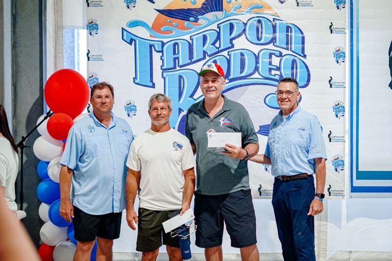 Four men pose in front of a Tarpon Rodeo sign, one holding a plaque, with red balloons nearby.