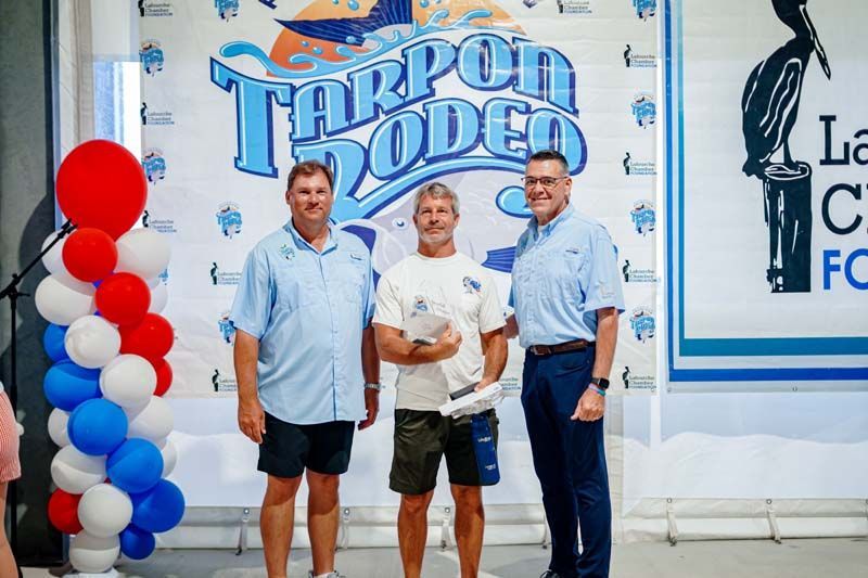 Three men posing at a Tarpon Rodeo event with red, white, and blue balloons beside them.