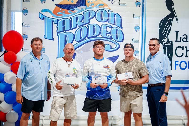 Five men hold certificates in front of a “Tarpon Rodeo” banner, with a fishing sculpture beside them.