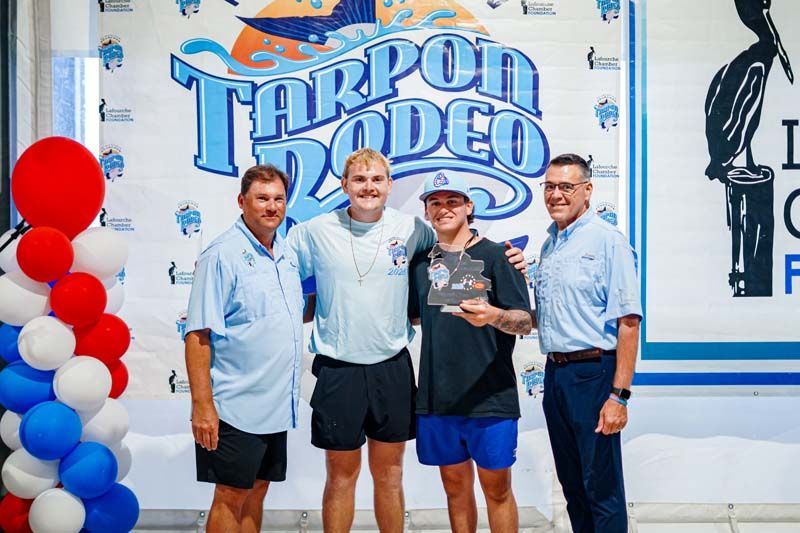 Four people posing in front of a Tarpon Rodeo sign, one holding a trophy, with red and white balloons nearby.