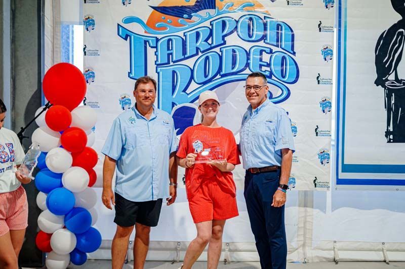 Three people posing in front of a “Tarpon Rodeo” sign, with red and white balloons nearby.